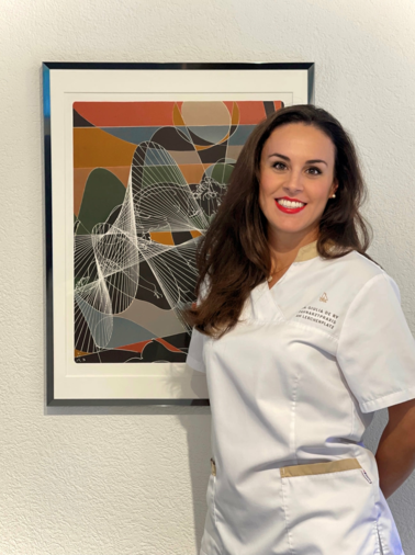 Portrait von Dr. med. dent. Giulia De Ry, die l&auml;chelnd in wei&szlig;er Praxiskleidung neben einem gerahmten, abstrakten Kunstwerk steht.